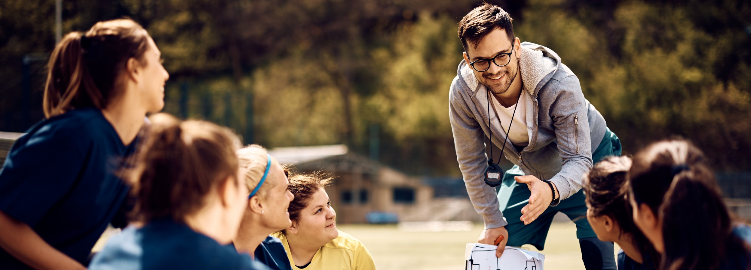 Ein Fußballtrainer erklärt die Spielstrategie auf einem sonnigen Fußballplatz den jungen Spielerinnen seiner Mannschaft.