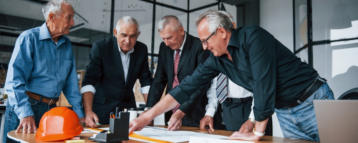 Eine Gruppe älterer Geschäftsmänner versammelt sich um einen Tisch, um einen Plan zu besprechen.  Das Bild zeigt fünf Männer mittleren Alters bis hohen Alters, die alle in einem geschäftlichen Umfeld stehen und sich über einen Plan beugen, der auf einem Tisch ausgebreitet liegt. Ein leuchtend oranger Bauhelm liegt am Rand des Tisches. Die Männer tragen eine Mischung aus formeller Kleidung, darunter Anzüge und Krawatten, sowie legerere Hemden und Jeans. Die Atmosphäre wirkt konzentriert und professionell, da alle auf das Dokument auf dem Tisch fokussiert sind. Das Büro scheint modern zu sein, mit großen Fensterflächen und einem sauberen, aufgeräumten Look. Die Stimmung ist ernsthaft und zielorientiert, was auf eine wichtige oder dringende Diskussion hindeutet.